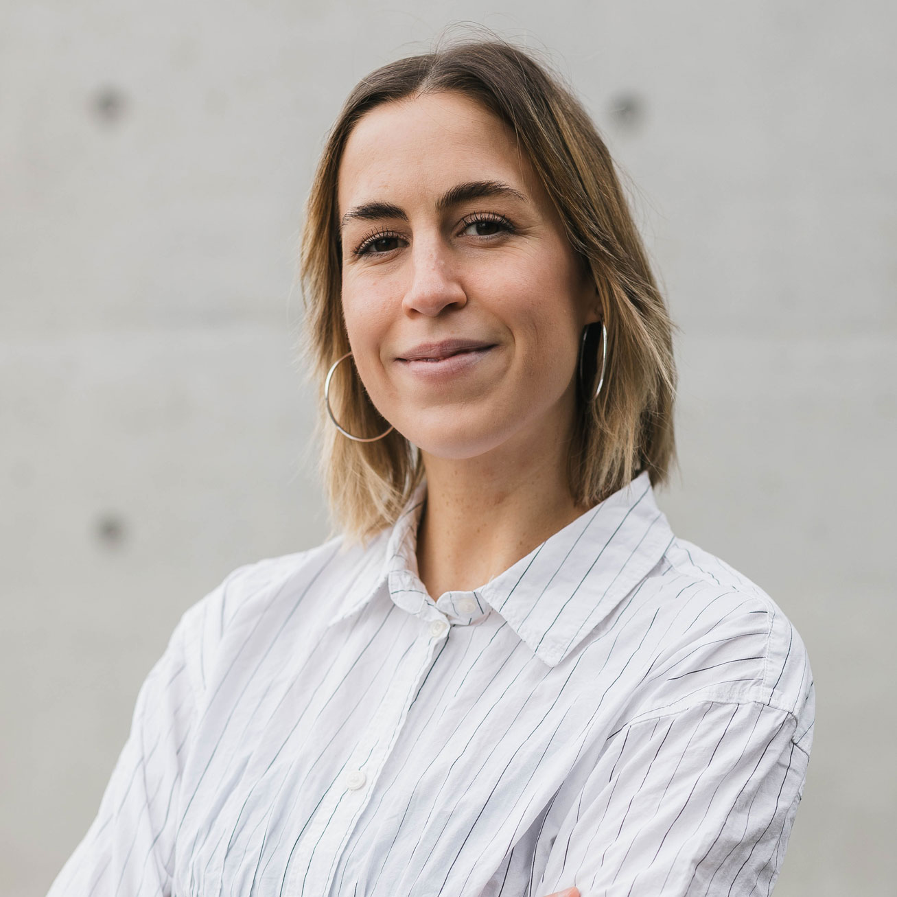 Porträt der Autorin Dorothea Winter. Sie steht mit verschränkten Armen vor einer hellgrauen Wand, trägt eine helle gestreifte Bluse und große Creolen und blickt mit leichtem Lächeln in die Kamera.